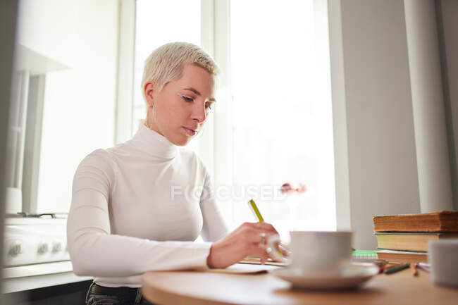 Lächelnde Astrologin macht sich Notizen im Notizblock am Schreibtisch mit einer Tasse Kaffee zu Hause im Sonnenlicht — Stockfoto