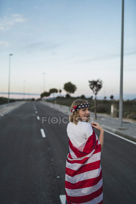Side view of delighted American female standing wrapped with national USA flag on roadway at sunset and looking away — Stock Photo