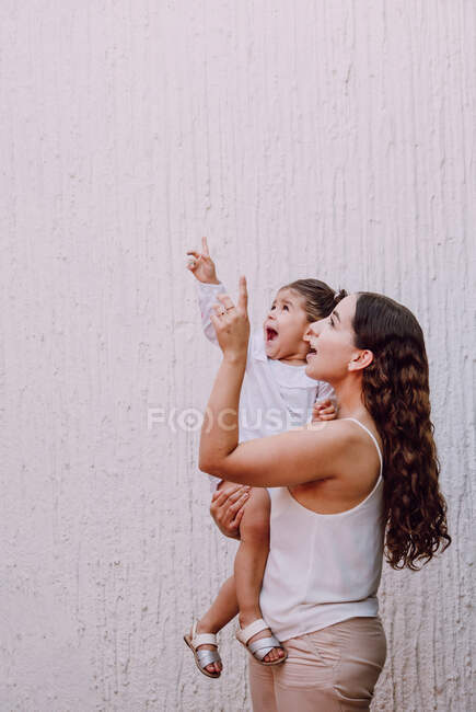 Vista lateral da mulher étnica de pé com a menina surpreendida na rua enquanto aponta e olha para cima — Fotografia de Stock