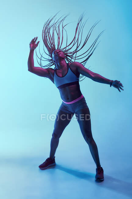 Strong African American female athlete in sportswear with flying Afro braids and biceps looking up in neon light — Stock Photo