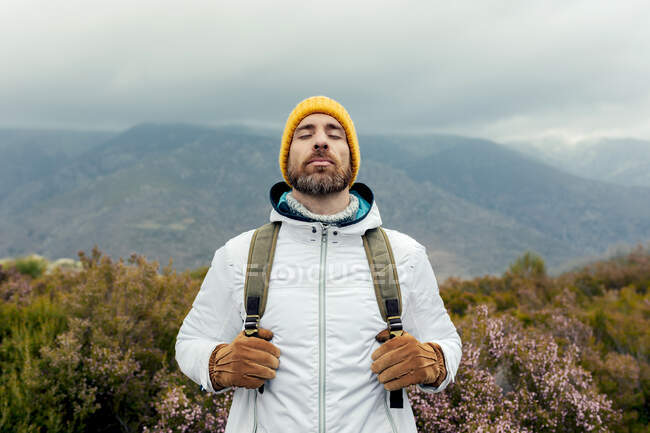 Восхитительный мужчина-кемпер в теплой шляпе, стоящий с рюкзаком в горах, наслаждающийся живописными видами с закрытыми глазами — стоковое фото