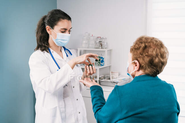 Female medic in uniform and disposable mask preparing medications for anonymous elderly patient on consultation in clinic during pandemic — Stock Photo