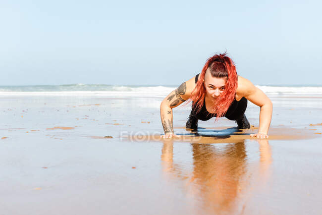 Визначена жінка-спортсменка з татуюваннями штовхає на піщане узбережжя, дивлячись на камеру під час тренувань проти океану — стокове фото
