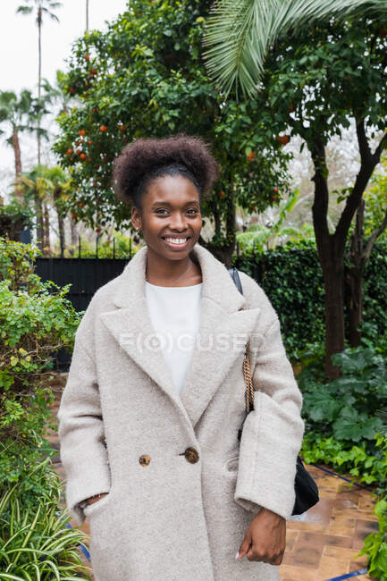 Gioiosa giovane donna afroamericana che indossa casual cappotto caldo in piedi con la mano in tasca nel verdeggiante parco cittadino e guardando la fotocamera — Foto stock
