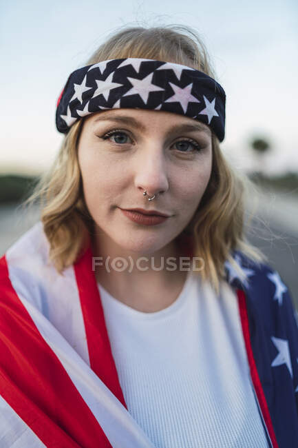 Retrato de encantadora mulher americana em bandana envolto em bandeira nacional dos EUA olhando para a câmera ao pôr do sol — Fotografia de Stock