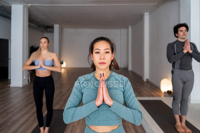 Серійна етнічна азіатка в спортивному одягу стоїть в Горі з молитовними руками позувати і робити йогу під час уроку групи в студії дивлячись на камеру — стокове фото