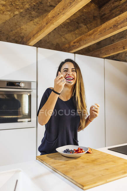 Candid weiblich mit Löffel Blick in die Kamera über Schüssel mit verschiedenen geschnittenen frischen Beeren in Schüssel zu Hause — Stockfoto