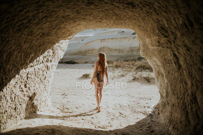 Back view unrecognizable fit female traveler wearing stylish swimsuit walking near stony natural cave in Sarakiniko Greece on sunny hot day — Stock Photo