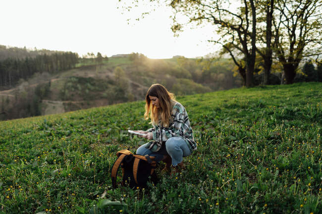 Молодая женщина турист наблюдает бумажный путеводитель во время приседания на лужайке против деревьев в солнечный день — стоковое фото