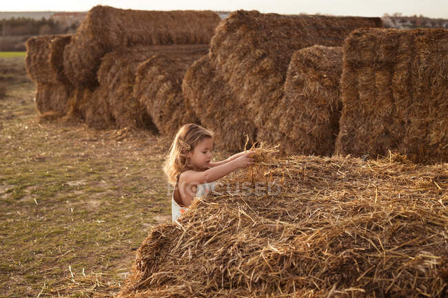 Side view of adorable child in overalls playing with hay near straw bales in countryside — Stock Photo