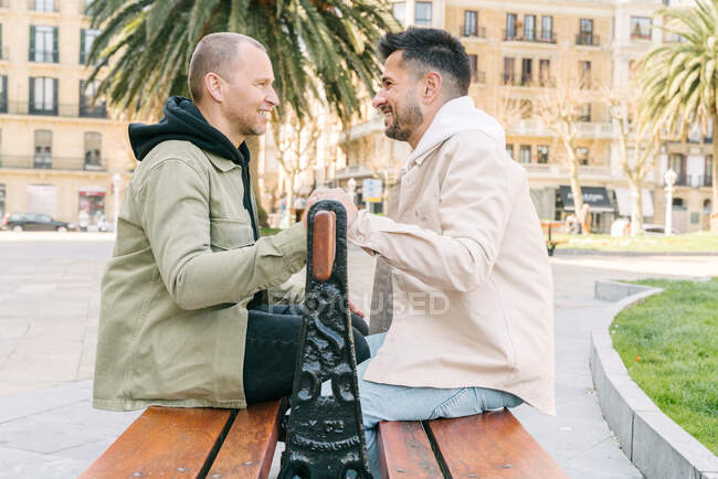 Side view of joyful young multiracial gay couple resting on wooden bench and looking at each other on city square on sunny day - foto de stock