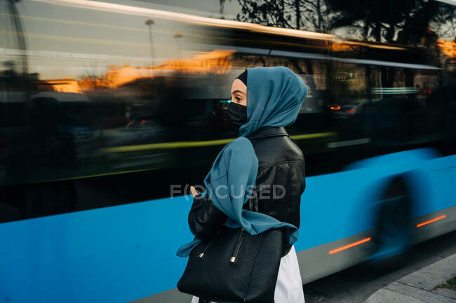 Vista trasera de la mujer musulmana en hijab de pie cerca de la carretera con tráfico ocupado en hora punta - foto de stock