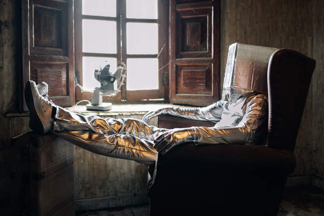 Side view unrecognizable person wearing protective silver suit with box on head resting on armchair in shabby room in abandoned house — Stock Photo