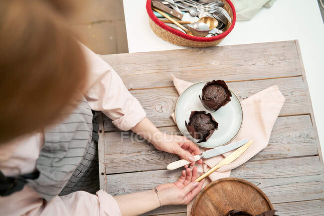 Сверху неузнаваемая женщина с ножами над вкусными кексами в чашках для выпечки на деревянном столе на кухне — стоковое фото