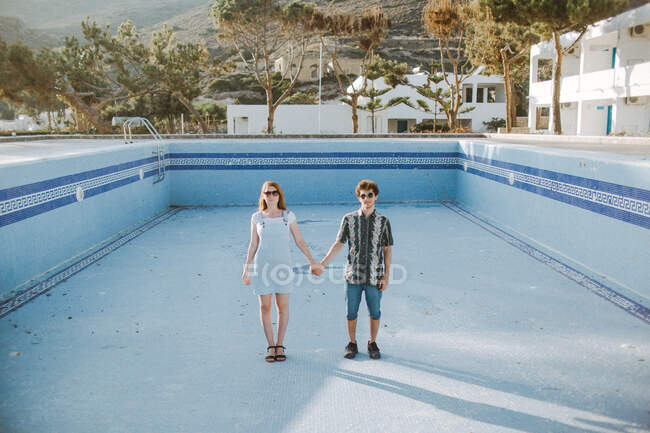 Ganzkörperjunges ruhiges Paar in lässigen Outfits, Händchen haltend und in die Kamera blickend, während es im Pool ohne Wasser im sonnigen Resort steht — Stockfoto