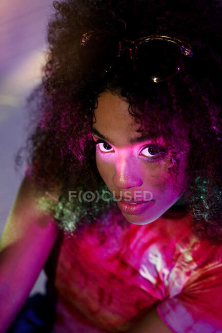 Dall'alto ritratto di attraente giovane donna afroamericana in abbigliamento casual guardando la fotocamera mentre in piedi in luci al neon — Foto stock