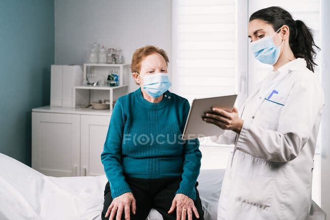 Médico femenino en uniforme con tableta que habla con una mujer mayor con máscara estéril en consulta durante la pandemia de coronavirus - foto de stock