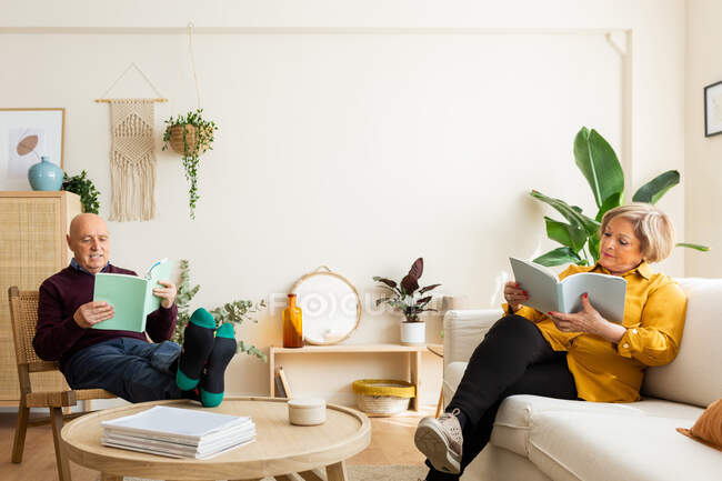 Casal de meia idade sentado na acolhedora sala de estar e lendo livros enquanto passa o tempo no fim de semana em casa — Fotografia de Stock