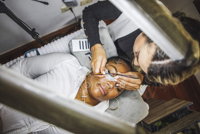 From above of female beauty master with tweezers applying fake eyelashes on face of ethnic client in salon — Stock Photo