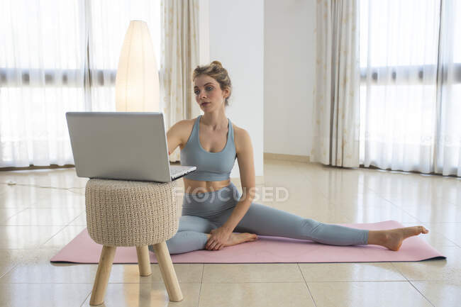 Tranquil hembra sentada en la esterilla y eligiendo video lección en línea en el portátil mientras se prepara para hacer yoga en casa - foto de stock
