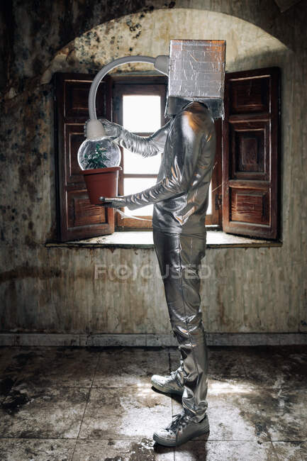 Vue latérale personne méconnaissable portant costume d'argent avec appareil respiratoire et tuyau fixé à la plante en pot debout dans la pièce abandonnée minable — Photo de stock