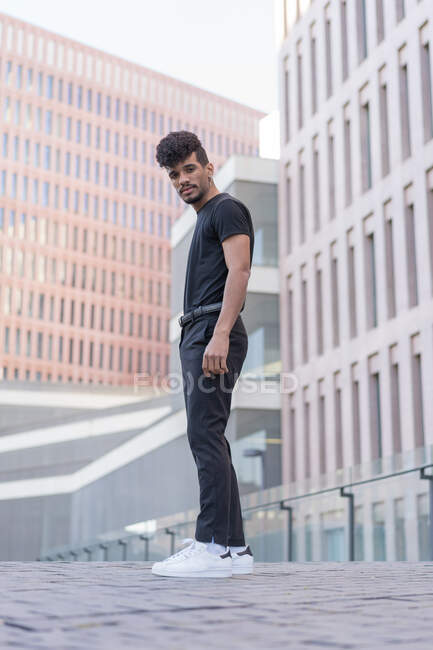 Ground level of confident ethnic african american male in black denim and t shirt standing on cobblestone pavement against contemporary buildings — Stock Photo