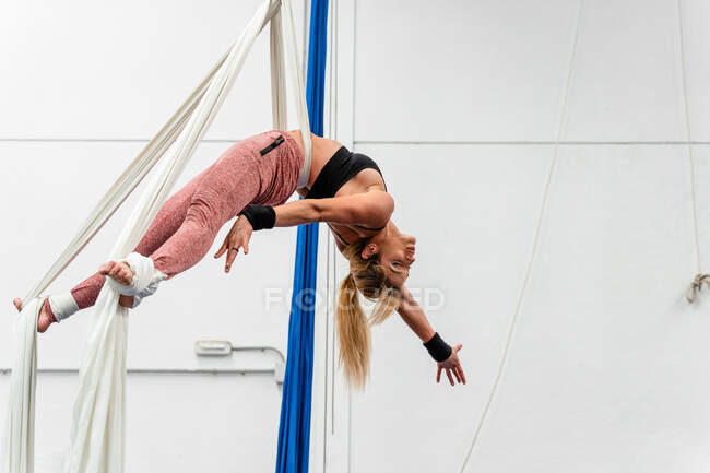 Полноразмерная гибкая спортсменка в активной одежде практикует воздушные упражнения на шелке в современном оборудованном спортивном клубе — стоковое фото