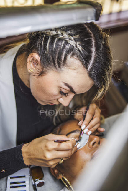 Female beauty master with tweezers applying fake eyelashes on face of ethnic client in salon — Fotografia de Stock