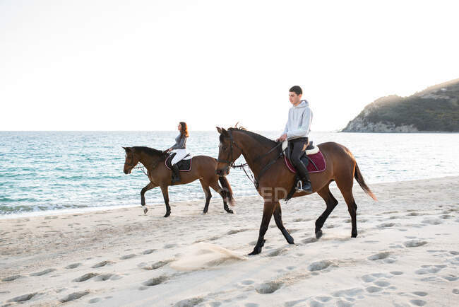 Side view of young woman with boyfriend riding purebred stallions on sandy shore against wavy ocean under blue sky — Stock Photo