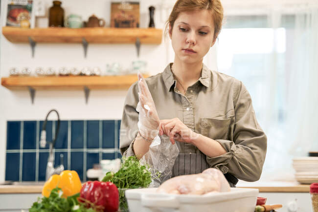 Crop female putting on transparent gloves and looking down against fresh peppers with herbs and raw chicken in baking pan at home — Stock Photo