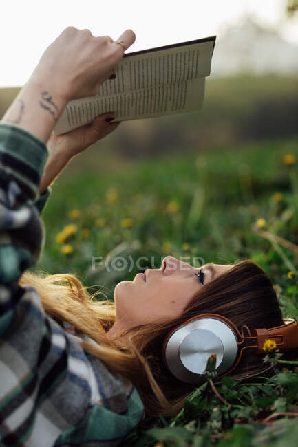 Side view of young attentive female in modern headset reading textbook while lying on meadow in summer — Fotografia de Stock