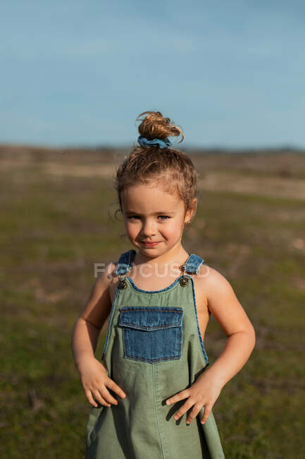 Delighted adorable little girl in overalls standing with hands on waist in meadow and looking at camera — Stock Photo
