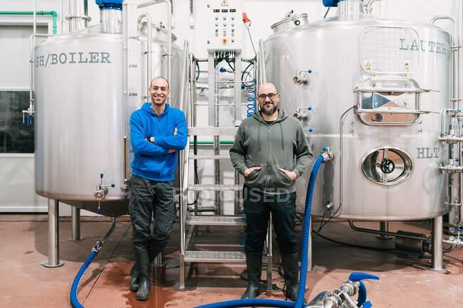 Коллеги-мужчины смотрят в камеру, стоя между металлическими цистернами в пивоваренной компании — стоковое фото
