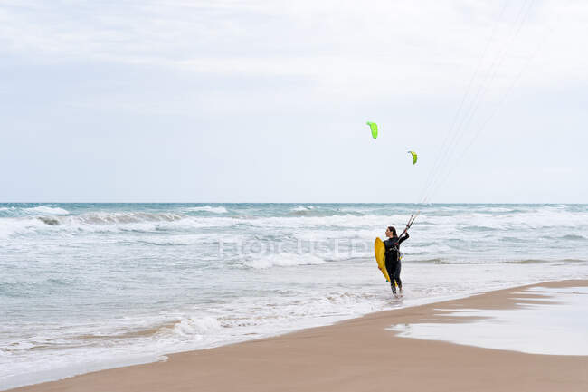 Atleta femenina en traje de neopreno con barra de control mirando hacia la orilla arenosa contra el océano espumoso después de practicar kitesurf - foto de stock