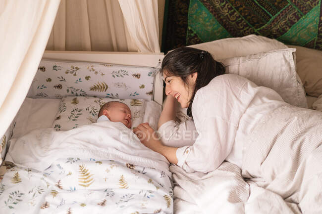 Счастливая мама держит за руки милого спящего новорожденного ребенка дома на размытом фоне — стоковое фото