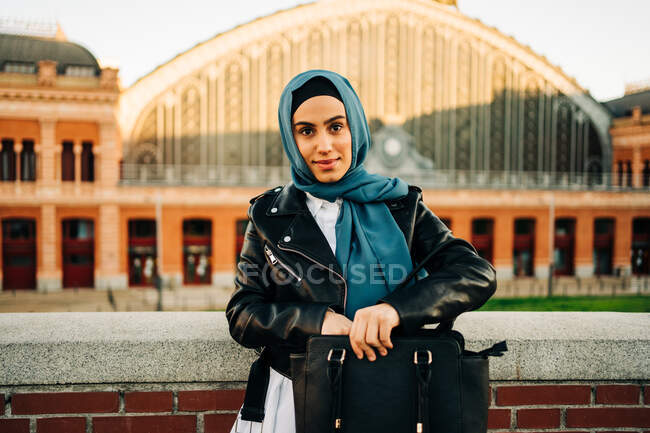 Stylish ethnic female in hijab standing on street and taking things out of leather handbag — Photo de stock