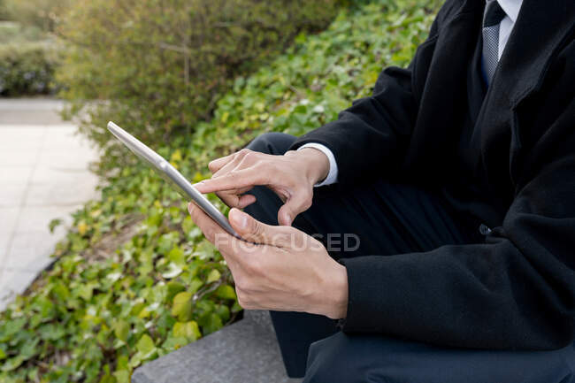 Cropped unrecognizable young well dressed ethnic male executive browsing internet on tablet while sitting on city in daylight — Stock Photo