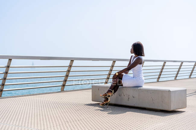 Seitenansicht in voller Länge trendige Afroamerikanerin in stilvollem weißen Kleid sitzt mit gekreuzten Beinen auf Steinbank an der Stadtpromenade und genießt Meerblick bei sonnigem Wetter — Stockfoto