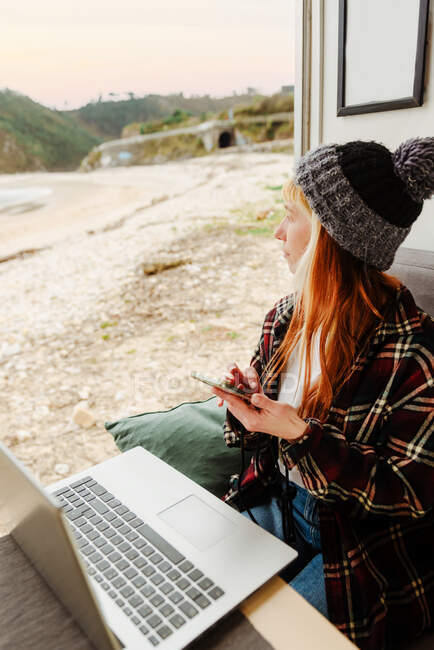 High angle of female traveling freelancer sitting in parked truck with smartphone and laptop and working on remote project while looking away — Stock Photo