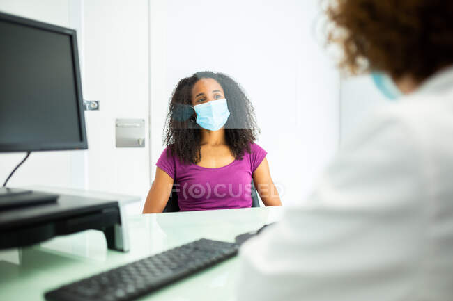 African American woman in face mask during appointment sitting at cropped unrecognizable female doctor's office desk protected by a perspex glass shield screen at modern clinic during coronavirus outbreak — Stock Photo