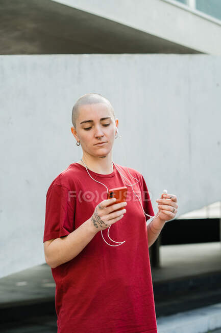 Молодая гомосексуальная женщина в футболке и наушниках с сотовым телефоном смотрит на экран, слушая музыку — стоковое фото