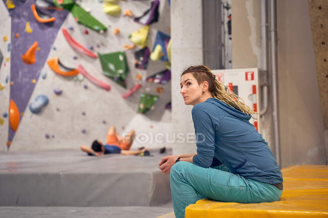 Vista lateral do montanhista profissional feminino sentado no tapete no moderno ginásio de escalada e olhando para longe — Fotografia de Stock