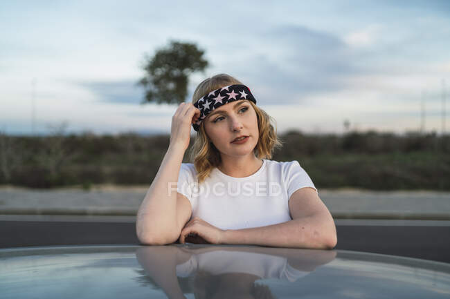 Giovane donna in abbigliamento casual e fascia con stampa bandiera americana appoggiata al finestrino dell'auto mentre si gode il viaggio al tramonto — Foto stock