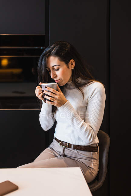 Conteúdo fêmea com caneca de bebida quente sentada no balcão na cozinha em casa e olhando para longe — Fotografia de Stock