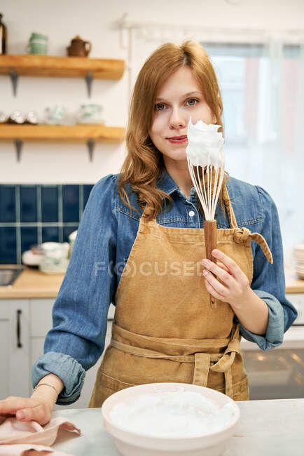 Улыбающаяся молодая женщина в фартуке со сладким кремом на венчике, смотрящая в камеру во время приготовления пищи дома — стоковое фото