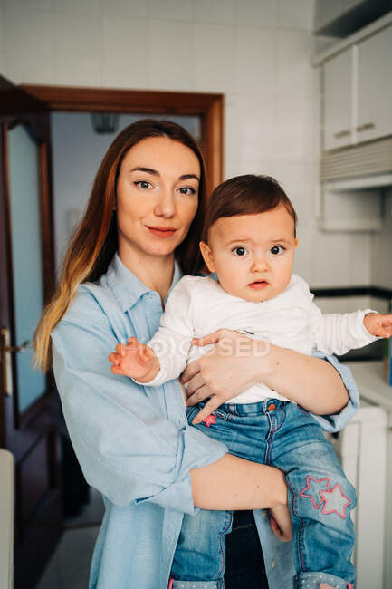 Giovane madre in abiti casual guardando la fotocamera mentre tiene piccolo bambino nella stanza della luce — Foto stock
