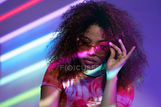 Ritratto di giovane donna afroamericana attraente in occhiali da sole alla moda guardando la fotocamera mentre in piedi in luci al neon — Foto stock
