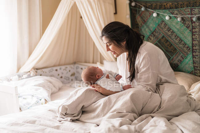 Улыбающаяся мама, взаимодействующая с неузнаваемым маленьким ребенком на измятой кровати дома при дневном свете — стоковое фото