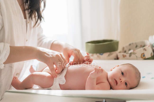 Crop anonymous mom putting diaper on charming little child looking at camera on infant table in house — Stock Photo