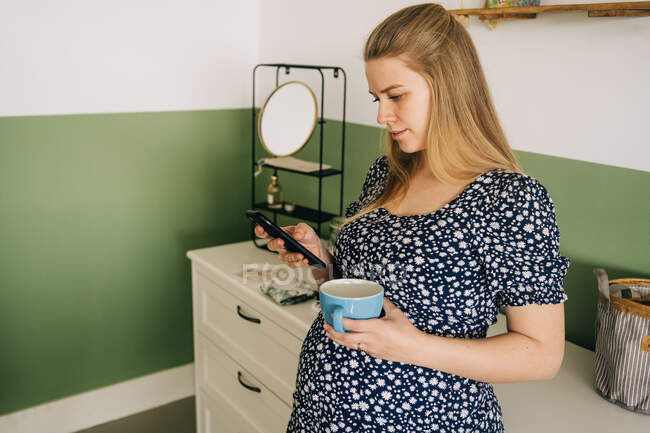 Giovane donna incinta in abbigliamento ornamentale con tazza di bevanda calda chatta sul cellulare in casa — Foto stock
